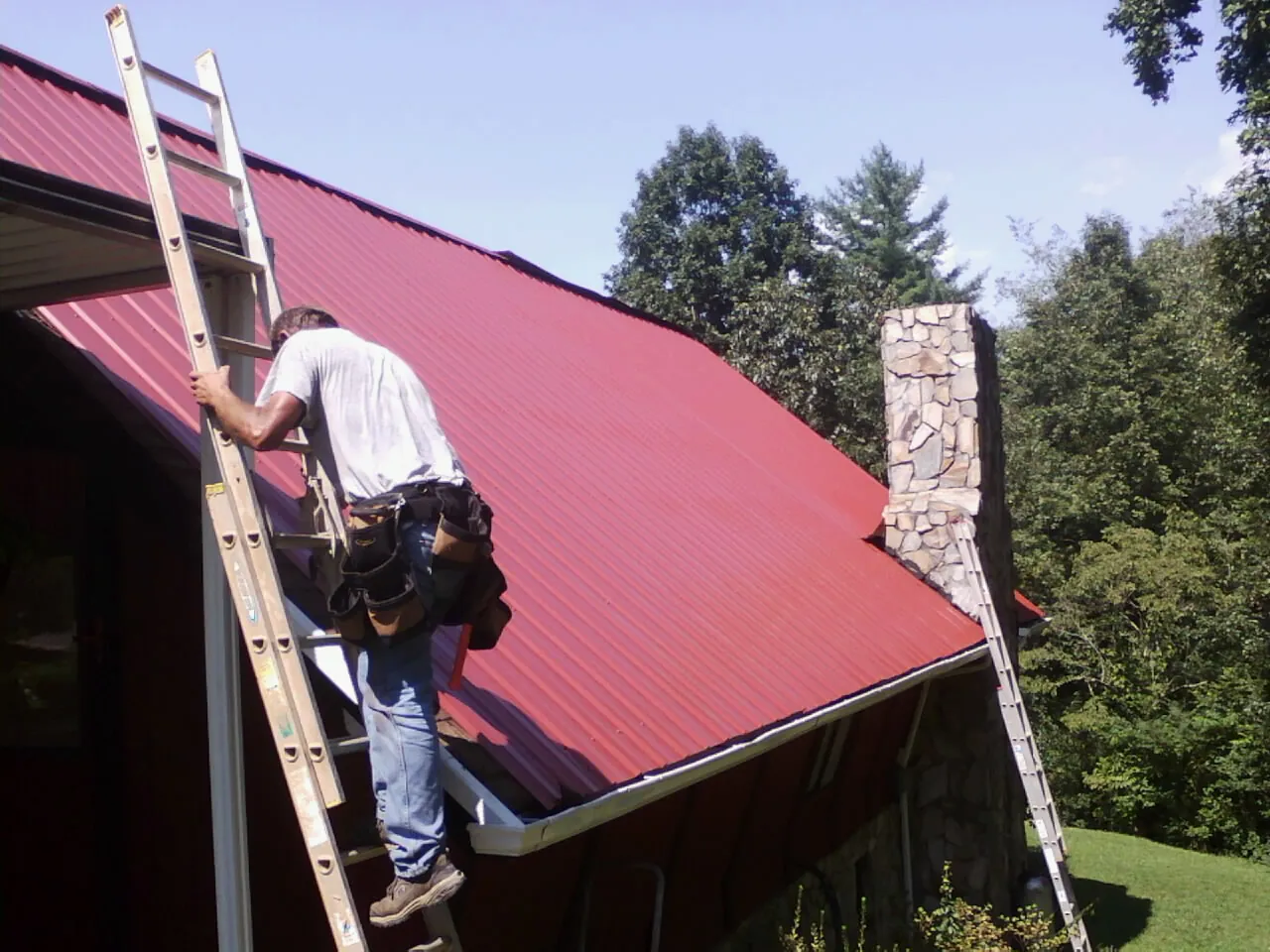 Roofer on a ladder beside a red metal roof and stone chimney — Soffit & Fascia Repair by Roofing Brothers in Springfield